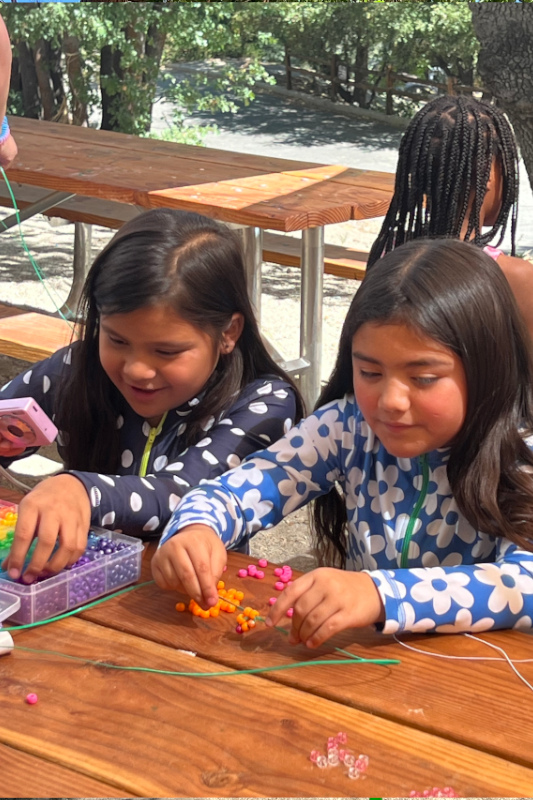 Two Hispanic young girl wearing making beaded bracelets at a picnic table.