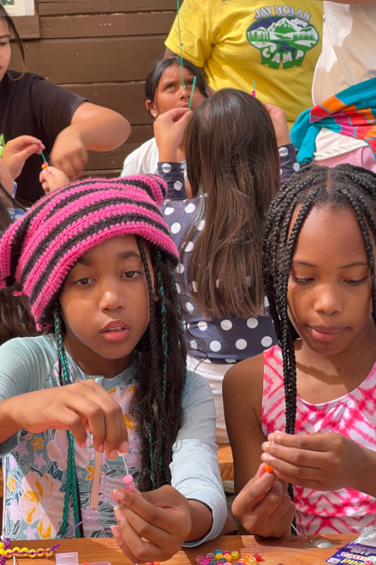 Two young female campers wearing colorful outfits making bead crafts together