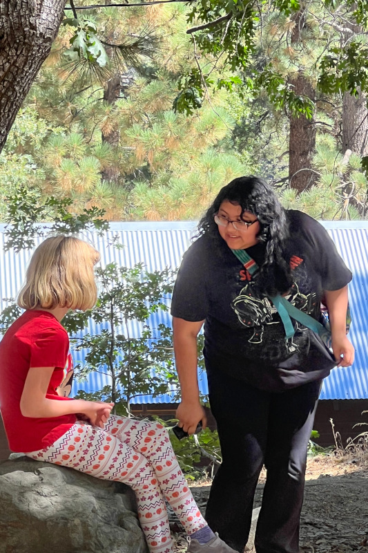 Young blond female camper sitting on a rock and talking with a female camp counselor dressed in black.