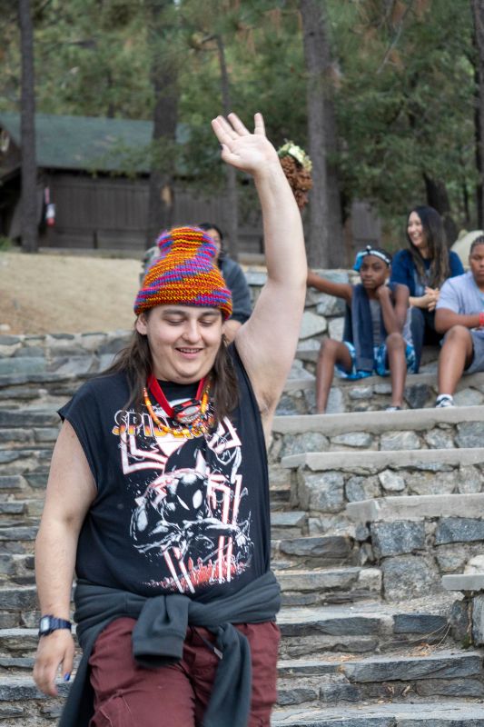 A man in a black graphic t-shirt and a colorful beanie holding his left hand up in the air while walking down the steps of an outdoor theater.