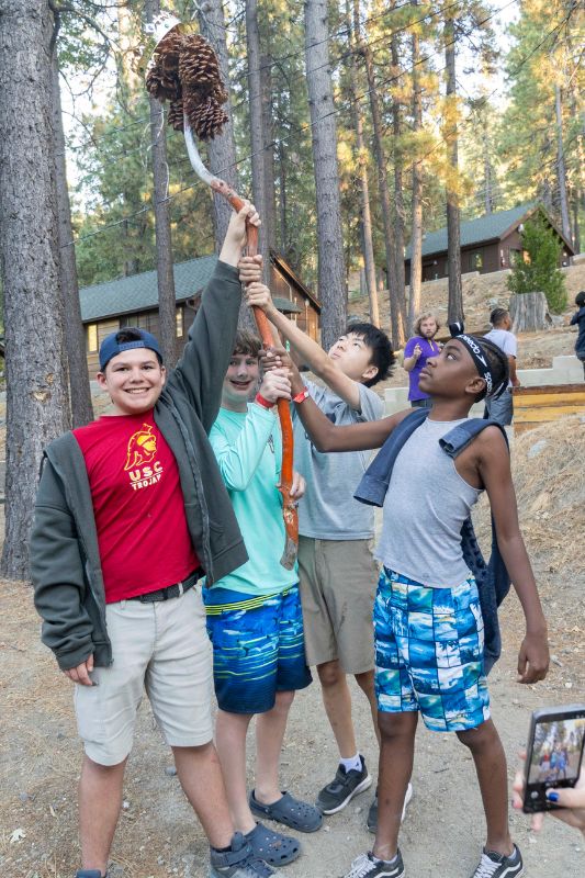 A group of male campers holding up a stick with pinecones attached to it.