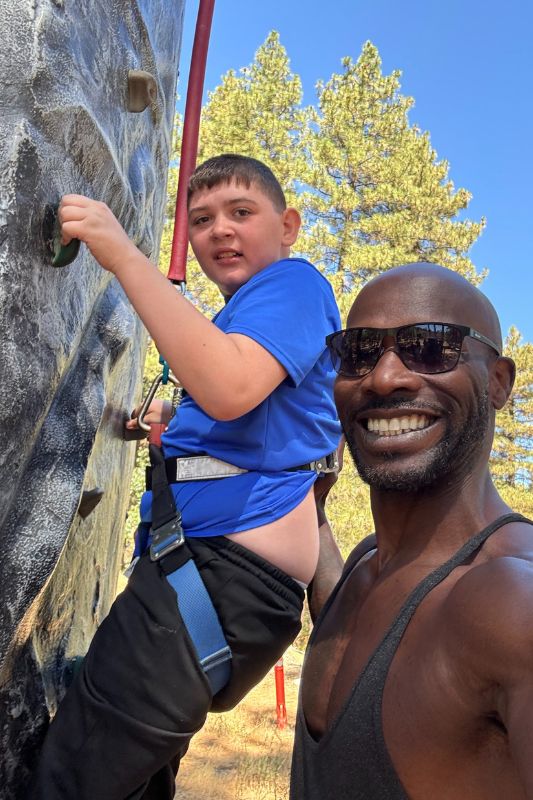 A kid in a blue shirt climbing a rock climbing wall with a counselor helping him