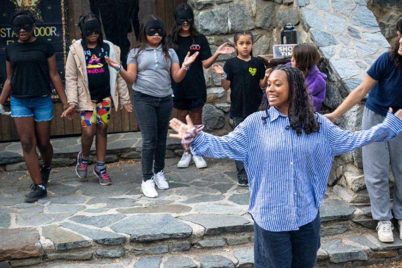 A female theater instructor teaching a drama class on a stone outdoor theater stage with students in the background